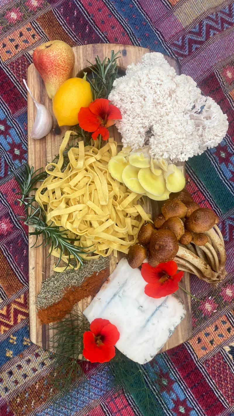 Pasta, mushrooms, blue cheese, a pear, a lemon, rosemary and nasturtium flowers laid out on a timber board over a woven rug