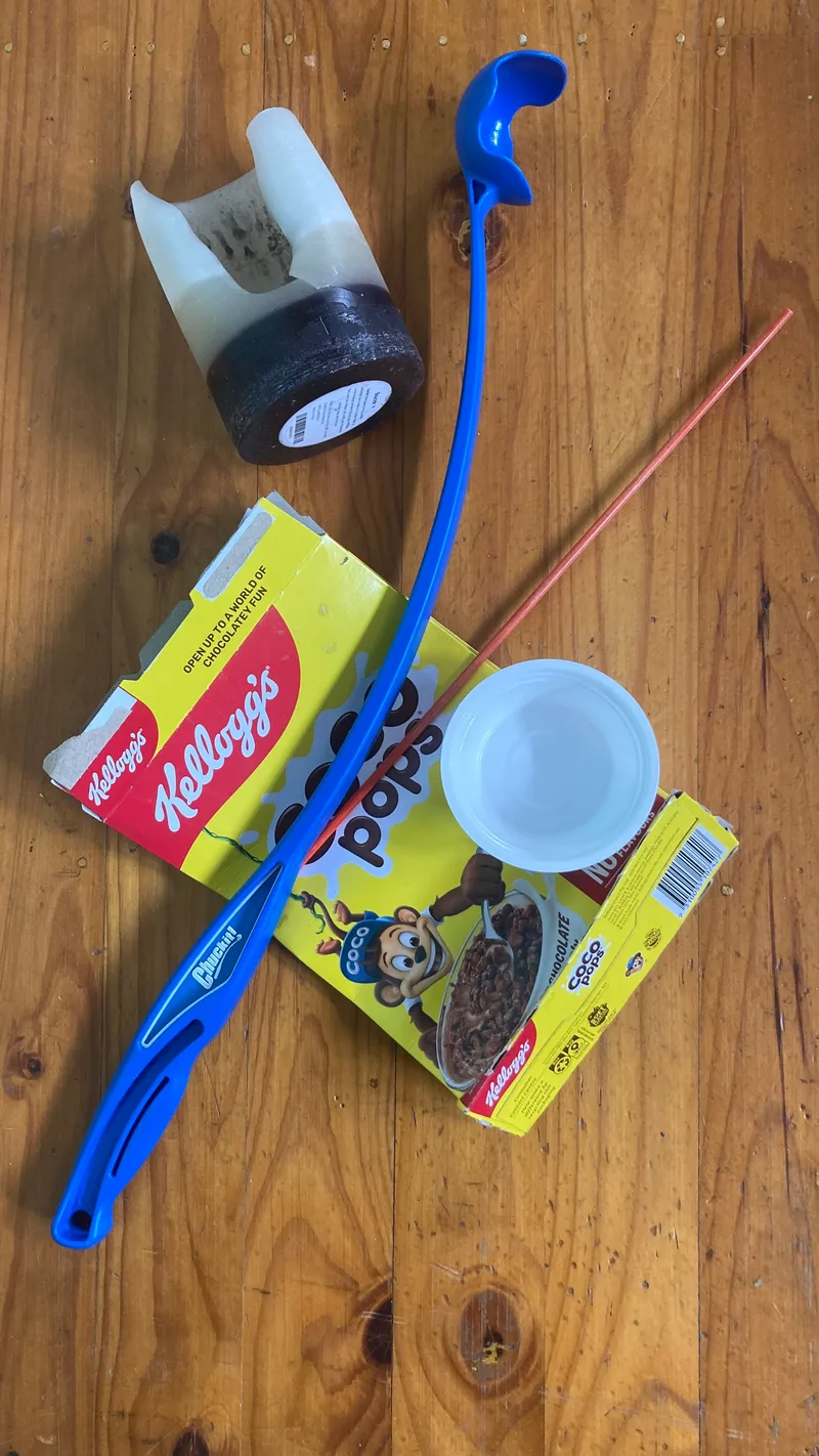 A cereal box, a blue ball thrower, a roll of tape, a plastic cup and a skewer on a timber table